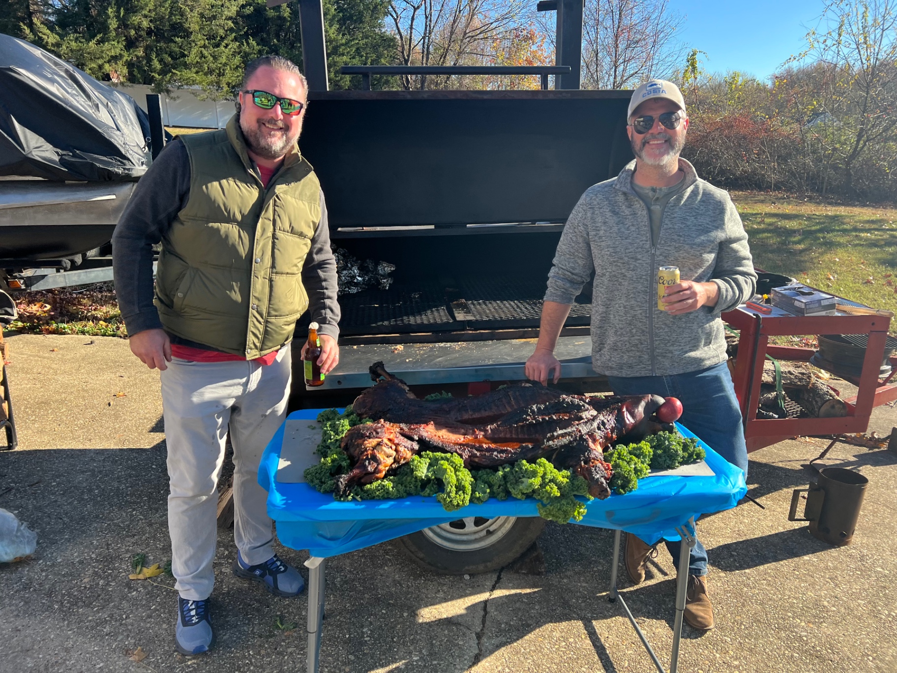 Two cooks standing behind a whole roasted hog beside a large smoker