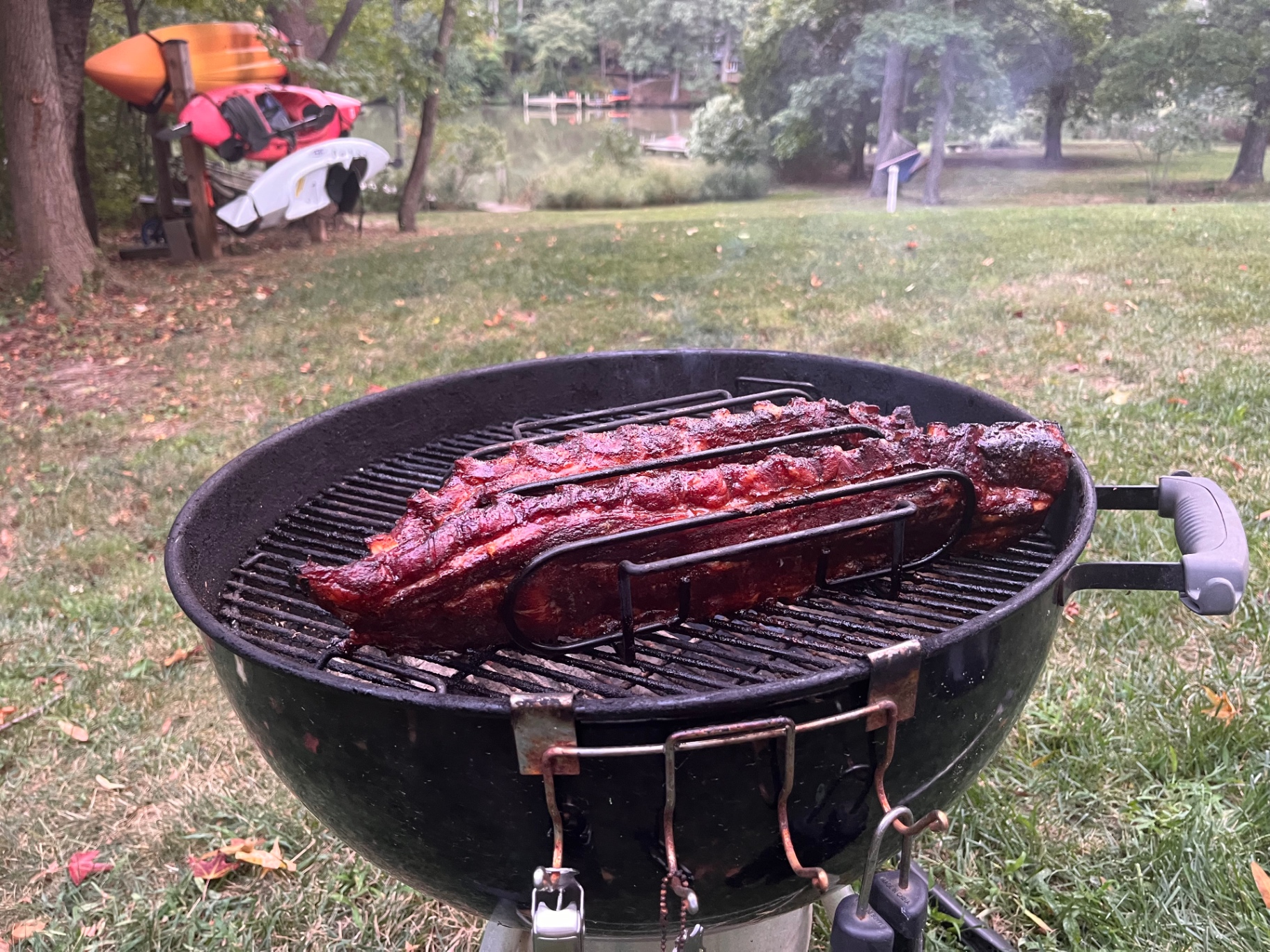 Two racks of pork ribs in a metal rib holder on a Weber kettle, waterfront in the background