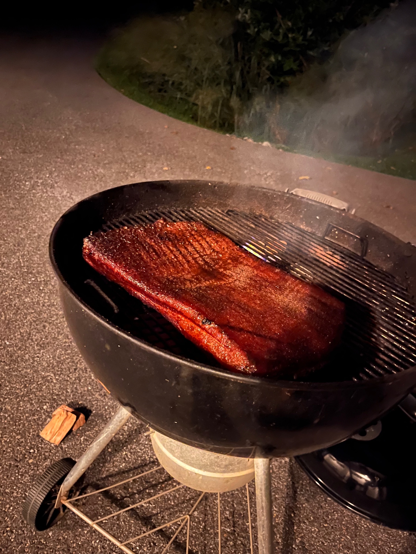 Pork belly darkening on the Weber kettle grate at night with a wood chunk nearby